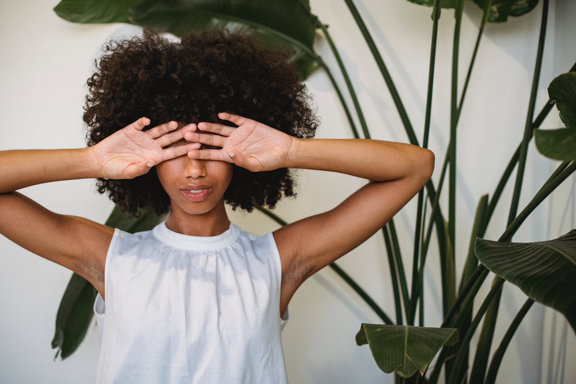 black woman with curls covering face with hands standing near potted plant