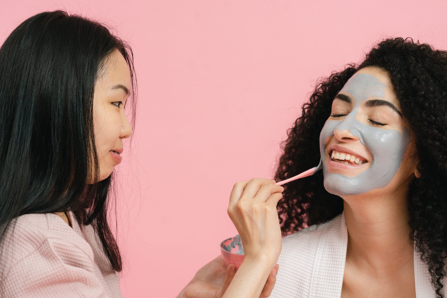 woman putting on cosmetic mask on face of other woman with curly hair